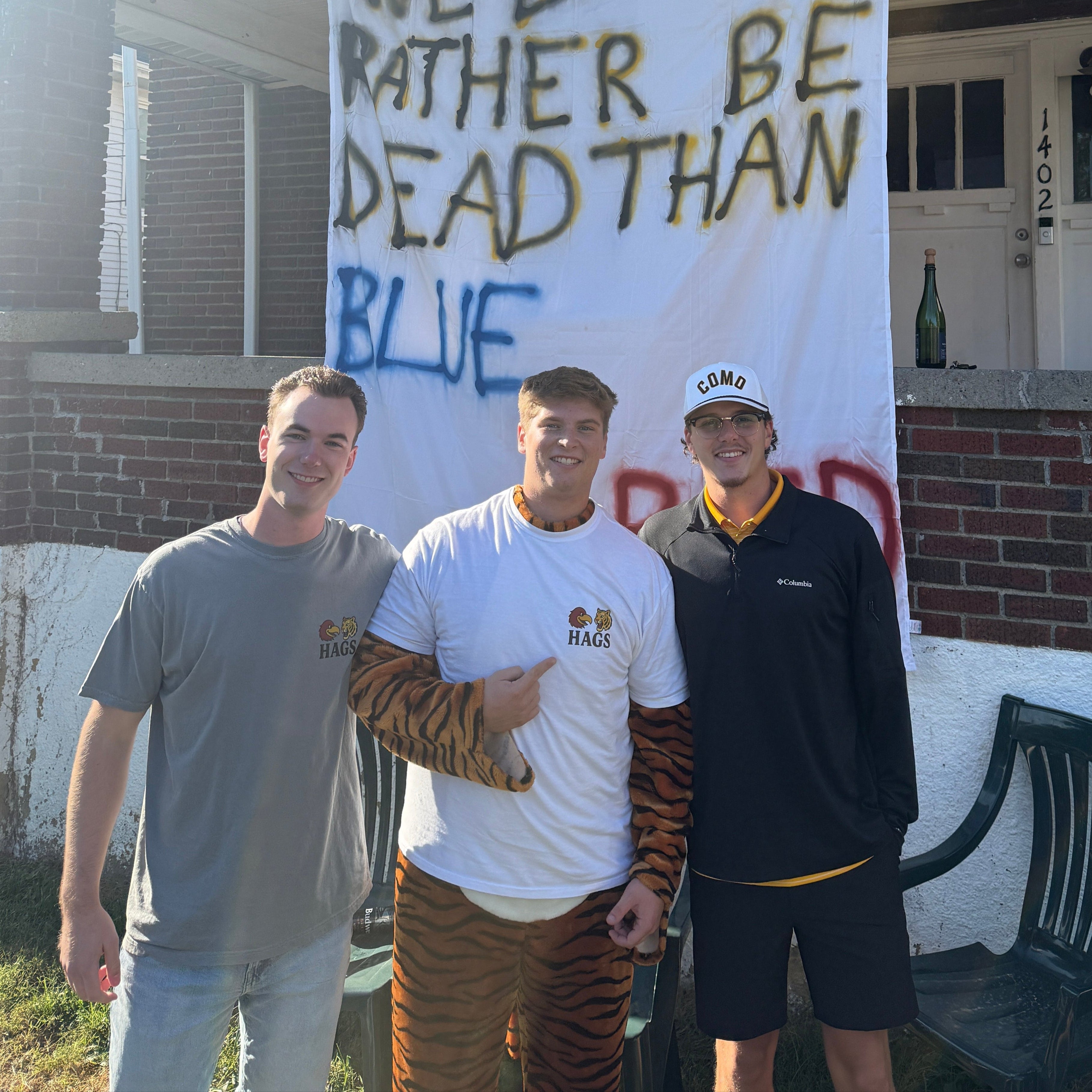 Two people standing in front of a house with a banner reading 'WE'D RATHER BE DEAD THAN BLUE'.