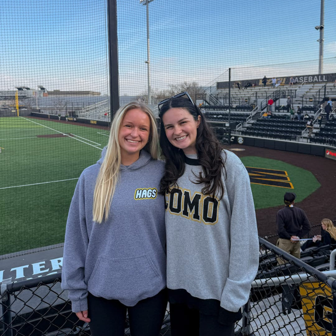 Two women standing on a baseball field with bleachers and stadium lights in the background.