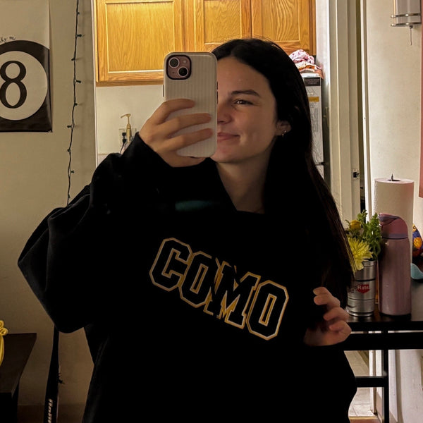 Person taking a mirror selfie wearing a black sweatshirt with 'COMO' text in a room.