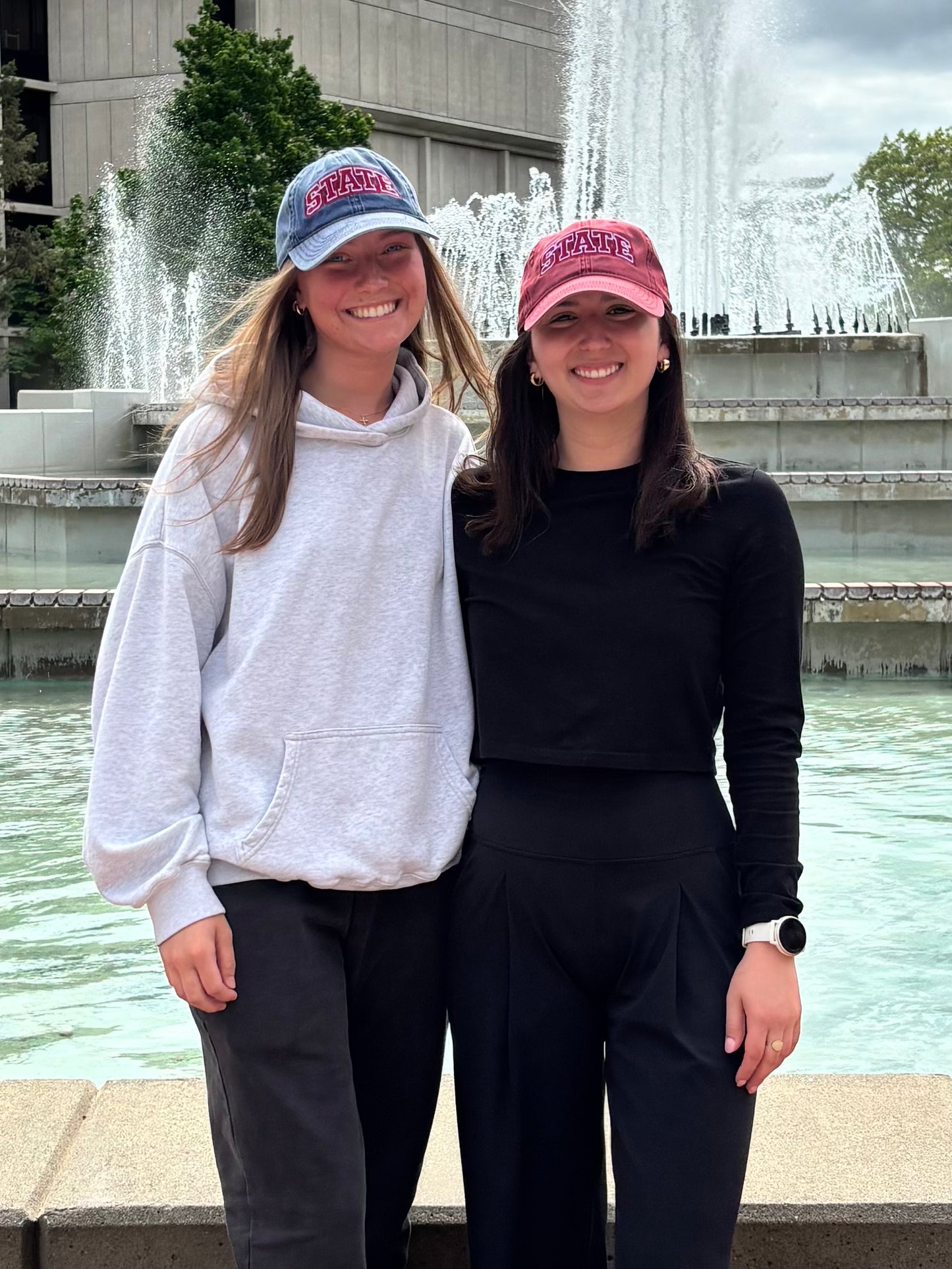 Two people wearing navy and burgundy HAGS STATE dad hats by a fountain.