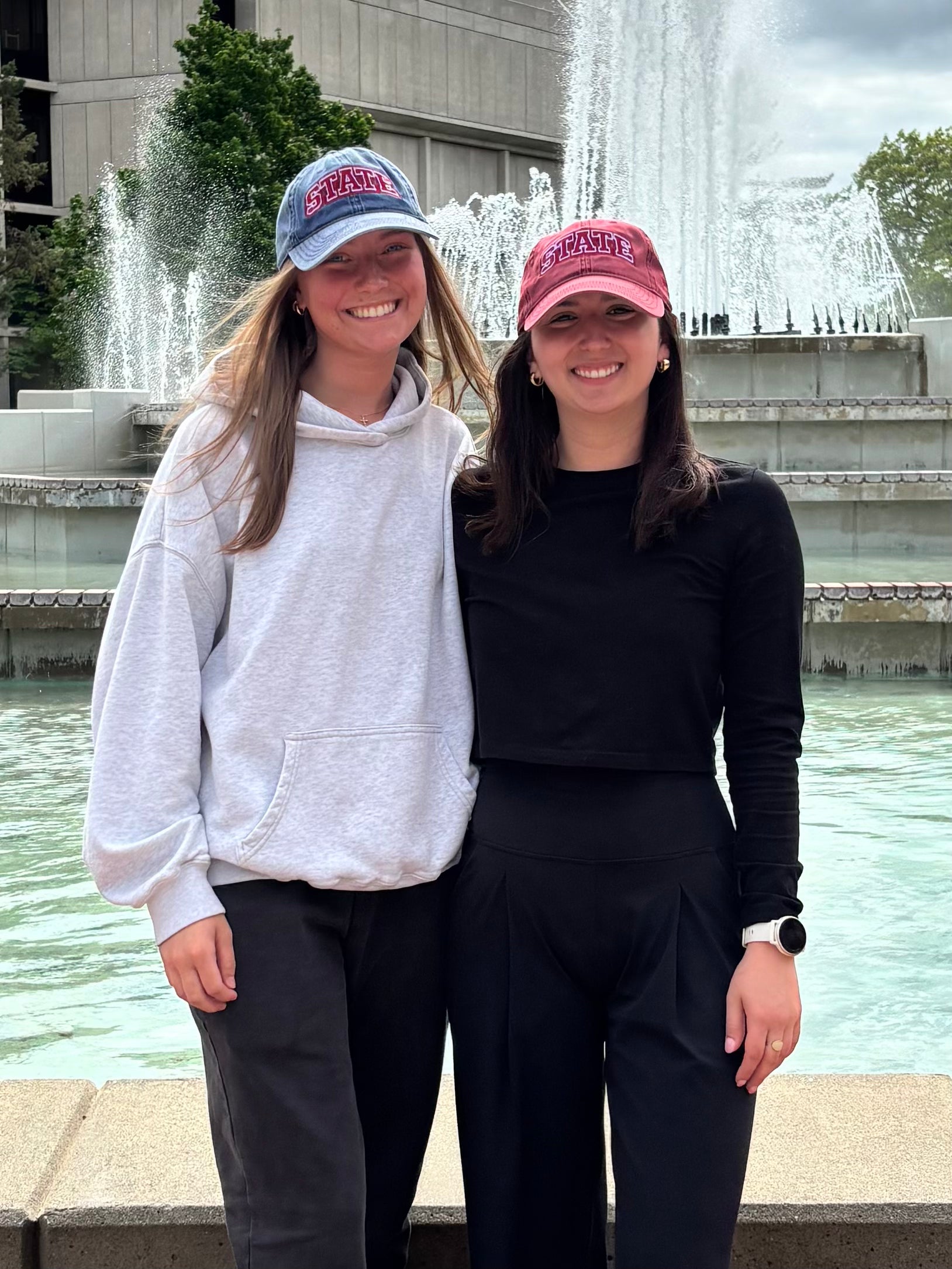 Two people wearing navy and burgundy HAGS STATE dad hats by a fountain.