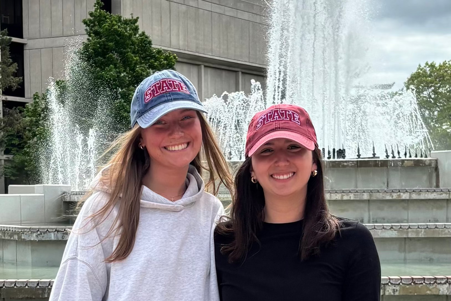 Two people wearing navy and burgundy HAGS STATE dad hats by a fountain.