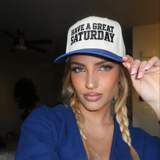 A woman wearing a white and blue trucker hat with the text 'HAVE A GREAT SATURDAY' in black, paired with a blue zip-up hoodie. She is taking a mirror selfie in an indoor setting.