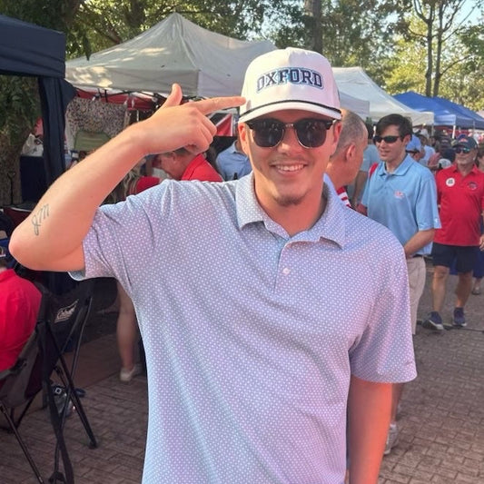 A man at an outdoor event wearing a white cap with 'EXPORB' printed on it, a light blue patterned polo shirt, and khaki shorts, making a shaka sign with his hand. The background shows a crowd, tents, and trees.